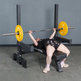 A person on the Bells of Steel Hydra Two Post Converter Bench grips a barbell with yellow plates, ready to lift in a gym with a gray wall, concrete floor, and various equipment attachments nearby.