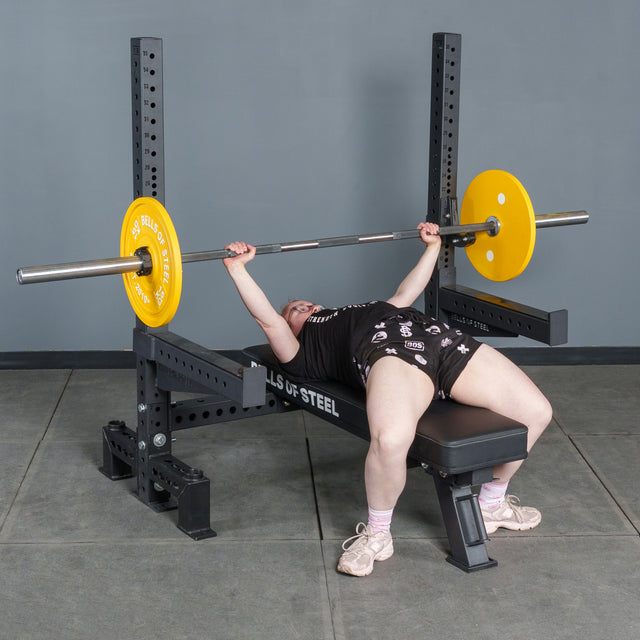 A person on the Bells of Steel Hydra Two Post Converter Bench grips a barbell with yellow plates, ready to lift in a gym with a gray wall, concrete floor, and various equipment attachments nearby.