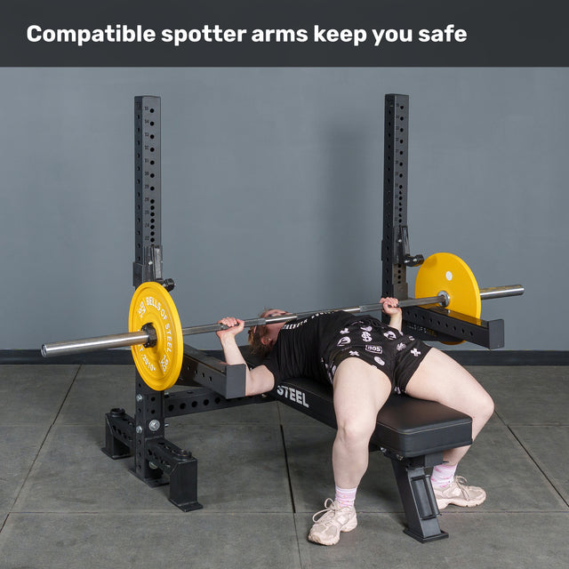 A person uses the Bells of Steel Hydra Two Post Converter Bench for a bench press with yellow weight plates. Text above reads, "Compatible spotter arms keep you safe.