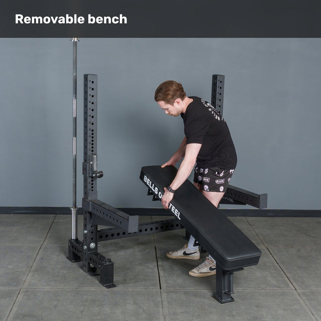 A man in a black shirt and shorts lifts the Bells of Steel Hydra Two Post Converter Bench from a weight rack in a gym. The text "Removable bench" is displayed at the top of the image.