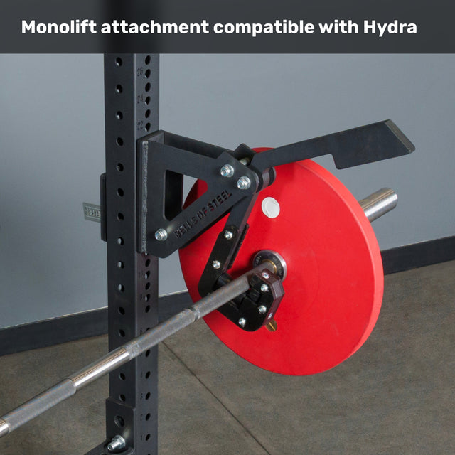 A close-up of a Bells of Steel Hydra Two Post Converter Bench with a monolift attachment on the squat rack, holding a barbell with a red plate. Text above: "Monolift attachment compatible with Hydra gym bench.