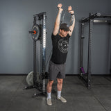 A man in a black cap, black t-shirt, and gray shorts uses the Bells of Steel Flat-Iron Tricep Press Cable Attachment to perform a standing overhead tricep extension in the gym, gripping the handle with both hands for optimal tricep engagement.
