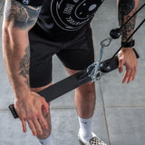 A tattooed person is using the Bells of Steel Flat-Iron Tricep Press Cable Attachment at the gym, gripping the black bar, while wearing a black t-shirt, black shorts, white socks, and checkered slip-on shoes.