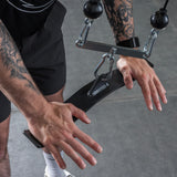 A person grips the Bells of Steel Flat-Iron Tricep Press Cable Attachment, focusing on tricep exercises. Their hands and the equipment are central, ready for activation, with the gym floor visible in the background.