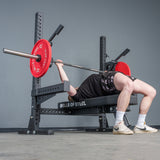A person in a black shirt and shorts, white socks, and sneakers uses the Bells of Steel Hydra Two Post Converter Bench, gripping a barbell with red plates in a gym.