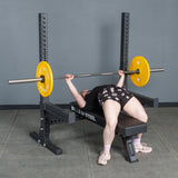 Wearing athletic clothing, a person bench presses a barbell with yellow plates on the Bells of Steel Hydra Two Post Converter Bench in a gym with gray walls and floor.