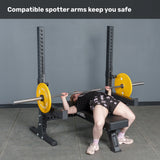 A person uses the Bells of Steel Hydra Two Post Converter Bench with yellow weight plates and safety spotter arms on a power rack. Text above: "Compatible spotter arms keep you safe." Perfect addition to any bench station.
