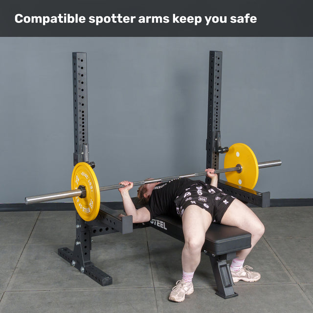 A person uses the Bells of Steel Hydra Two Post Converter Bench with yellow weight plates and safety spotter arms on a power rack. Text above: "Compatible spotter arms keep you safe." Perfect addition to any bench station.
