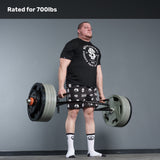 A man in a black t-shirt, patterned shorts, and socks lifts a heavily loaded Bells of Steel Open Trap Bar / Hex Bar in a gym. Text above reads "Rated for 700lbs." The background is plain gray.