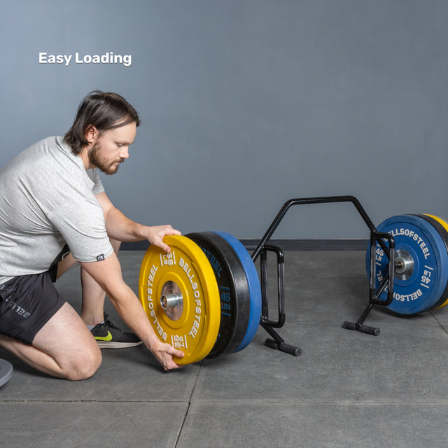 A man kneels, sliding a yellow plate onto a Bells of Steel Open Trap Bar loaded with blue and black weights. "Easy Loading" is shown on the wall, highlighting the hex bar’s rotating sleeves for quick plate changes.