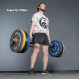 A man lifts the Bells of Steel Open Trap Bar / Hex Bar, rated for 700lbs, in a gym. He wears a grey t-shirt and black shorts. The bar features rotating sleeves; the background is a plain gray wall and floor.