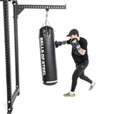 A person in a black T-shirt, cap, and sweatpants lands powerful punches on a Bells of Steel Heavy Bag Set. This set includes a robust metal frame securely mounting the punching bag against a plain white background, capturing the essence of combat sports.