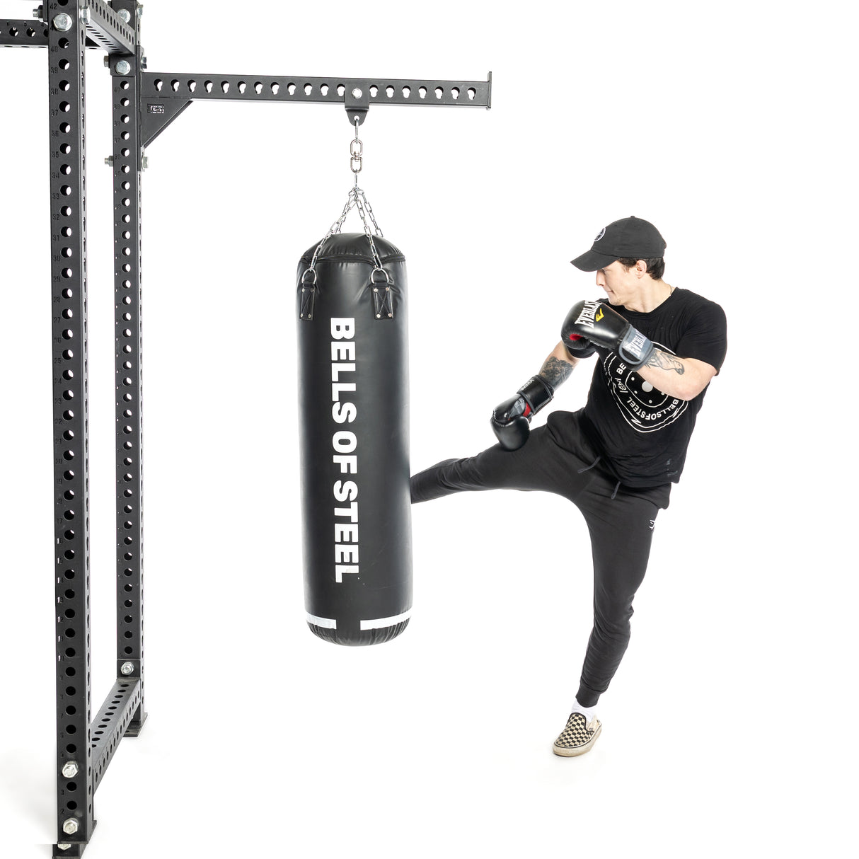 Dressed in black athletic gear and a cap, the individual delivers a powerful kick to a hanging punching bag. The Bells of Steel Heavy Bag Set sways under the blow, while boxing gloves and checkered shoes enhance their dynamic movement, reflecting true combat sports training spirit.