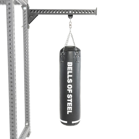 A Bells of Steel Heavy Bag Set in black hangs from a chain attached to a metal Power Rack with holes, displayed against a plain white background.