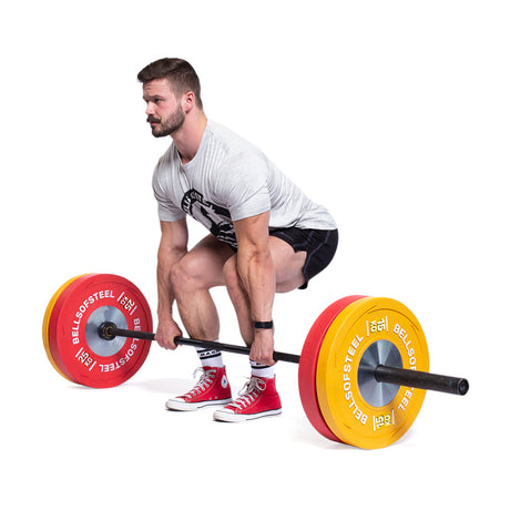 A man in shorts, a t-shirt, and red sneakers prepares to lift a barbell loaded with Bells of Steel KG Competition Bumper Plates, built to IWF specs. He is gripping the bar with both hands and bending at the knees.