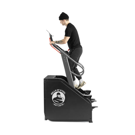 A person in a black shirt, pants, and beanie uses the Bells of Steel Sisyphean Stepper - Manual Stair Climber on a white background, demonstrating an effective cardio workout with adjustable resistance.