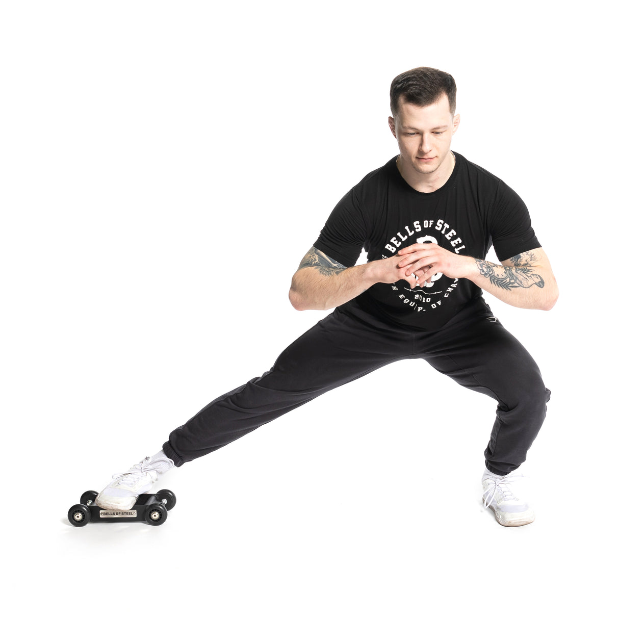 A tattooed man in a black Bells of Steel T-shirt and pants performs a lateral lunge using a Glute Ham Slider under his right foot, engaging his posterior chain. He balances with both arms forward against a white background.