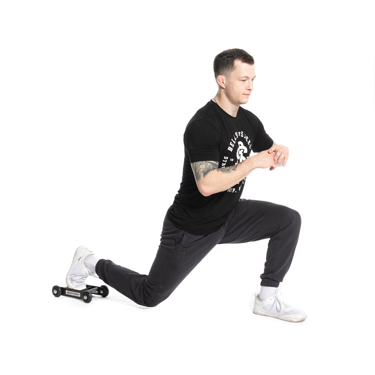 A person in a black t-shirt and pants is kneeling in a lunge position on a white background, with one knee on the Bells of Steel Glute Ham Slider and hands clasped in front.