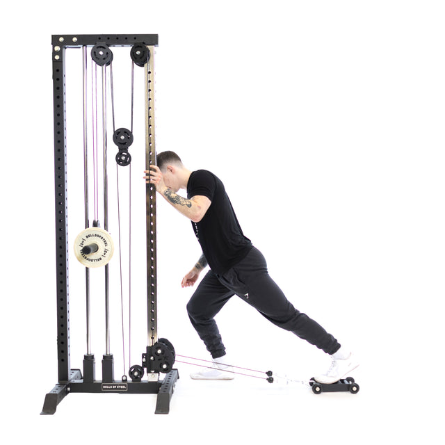 Using the Glute Ham Slider by Bells of Steel, a person is working on their posterior chain with resistance bands on their ankles. They lean forward, extending one leg back while wearing a black outfit and white sneakers.