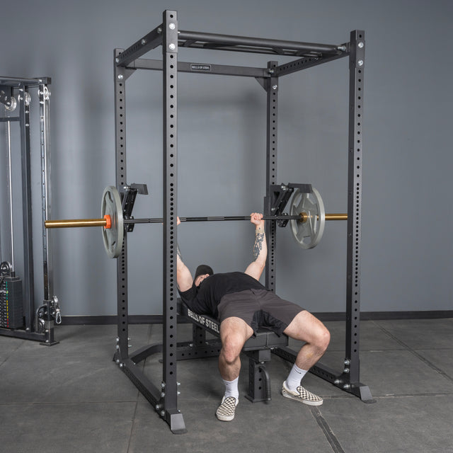 A person performs a bench press on a bench inside a power rack, using the Bells of Steel Monolift Rack Attachment to hold the barbell above their chest.