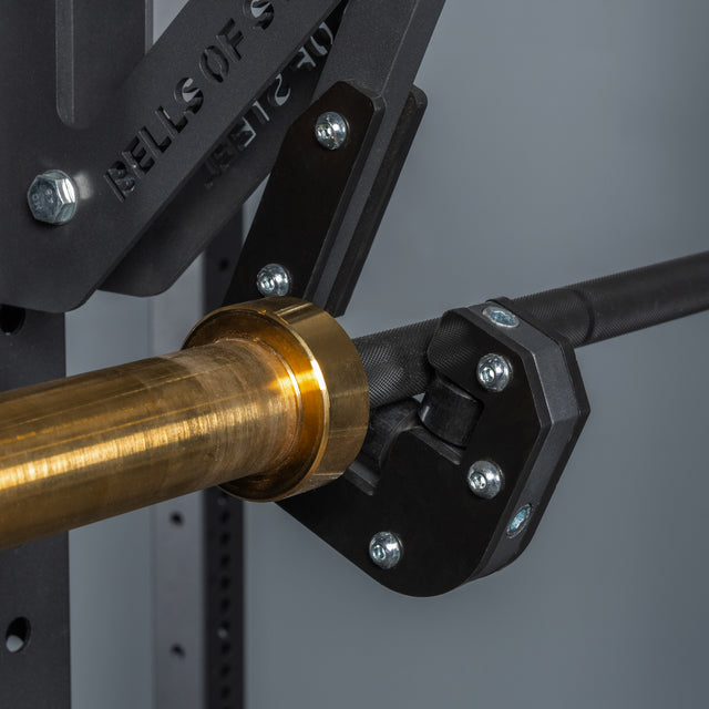 Close-up of a Bells of Steel Monolift Rack Attachment holding a metallic barbell with a gold sleeve, mounted on a black metal frame with visible bolts and engraved text.