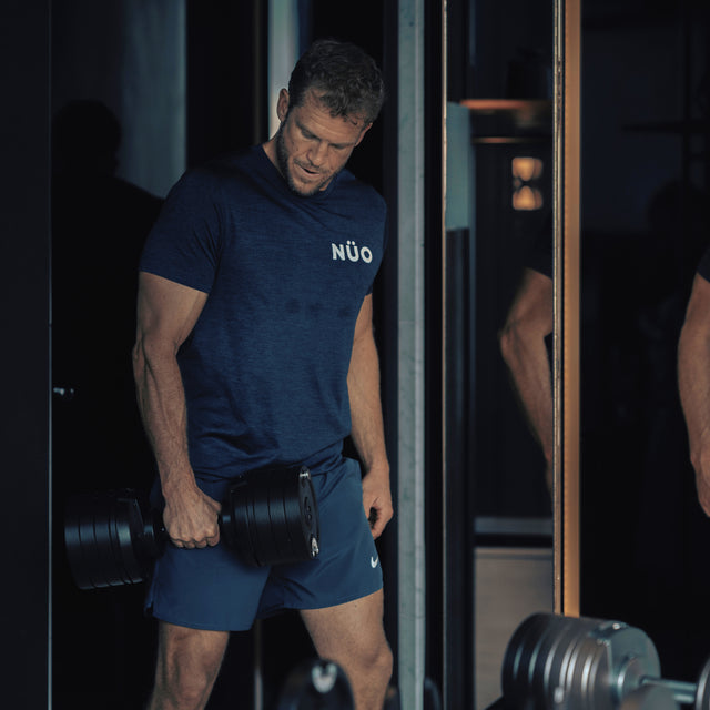 A man in a blue NUO t-shirt and shorts stands indoors, gripping a Bells of Steel NÜOBELL-S Adjustable Dumbbell with focused intensity in a gym or fitness studio.