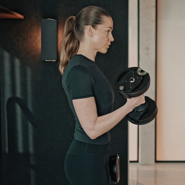 A woman in athletic wear stands in profile, performing a bicep curl with a Bells of Steel NÜOBELL-S Adjustable Dumbbell, highlighting strength and focus in a sleek modern home gym setting.