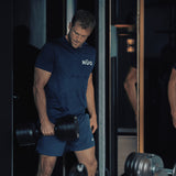 A man in a navy blue shirt lifts Bells of Steel NÜOBELL-S Adjustable Dumbbells in a gym, standing before a mirror. Focused and muscular, he shows sweat stains on his shirt while using the precision-engineered equipment.