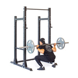 A person in black workout gear performs a squat with a barbell, using the sturdy Bells of Steel Hydra Half Rack (3" x 3", ⅝" Holes) for support. The plain white floor highlights the sleek gym setup, ideal for home fitness enthusiasts.