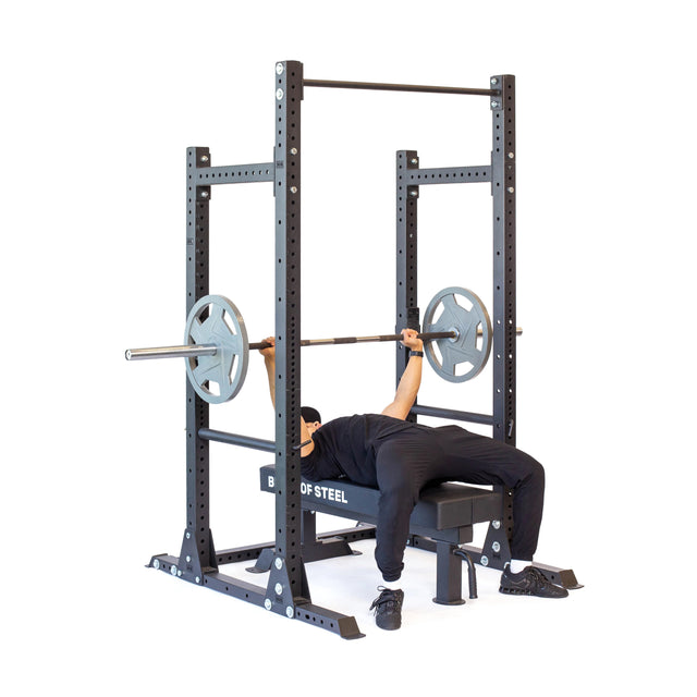 A person in black workout clothes performs a bench press on the robust Hydra Half Rack - Prebuilt (3" x 3", ⅝" Holes) by Bells of Steel, ideal for a home gym. The barbell is loaded with weights against a plain white background, showcasing the rack's rugged elegance.