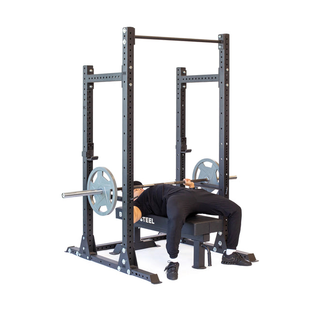 A person, dressed in athletic gear and comfortably positioned in their home gym with feet flat on the ground, is lying under a loaded barbell on a Hydra Half Rack - Prebuilt (3" x 3", ⅝" Holes) by Bells of Steel, ready to execute a bench press.