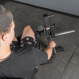 A person in a black shirt, printed shorts, and a cap uses a seated calf raise machine next to the Bells of Steel Legacy Leg Extension / Hamstring Curl Machine - Plate Loaded in a gym with gray flooring.