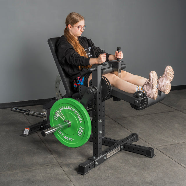 A person with long hair and glasses uses the Bells of Steel Legacy Leg Extension / Hamstring Curl Machine - Plate Loaded in a home gym. The machine has a green weight plate and sits on a gray floor. The user wears a black hoodie, shorts, and pink sneakers.