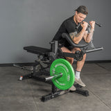 Wearing athletic wear, a person does preacher curls with dumbbells on a black and green bench in a gym with gray walls, next to the Bells of Steel Legacy Leg Extension / Hamstring Curl Machine - Plate Loaded.
