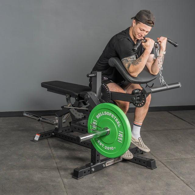 Wearing athletic wear, a person does preacher curls with dumbbells on a black and green bench in a gym with gray walls, next to the Bells of Steel Legacy Leg Extension / Hamstring Curl Machine - Plate Loaded.