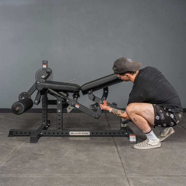 A person in a cap and shorts adjusts the settings of a black Bells of Steel Legacy Leg Extension / Hamstring Curl Machine - Plate Loaded in a home gym with gray walls and a concrete floor.