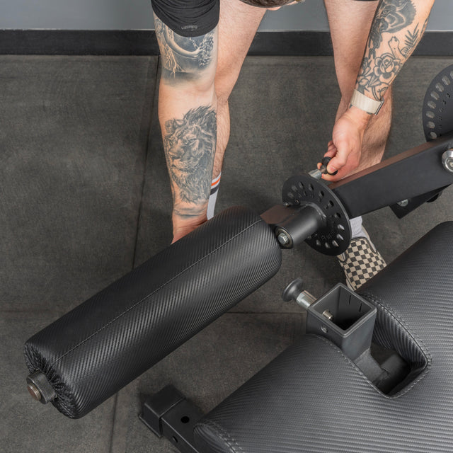 A person wearing checkered slip-on shoes adjusts the padded roller on the Bells of Steel Legacy Leg Extension / Hamstring Curl Machine - Plate Loaded. Only their tattooed legs and hands are visible.