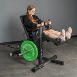 A woman in athletic wear uses the Bells of Steel Legacy Leg Extension / Hamstring Curl Machine - Plate Loaded with green weights in a gym, extending her legs—perfect for enhancing any home gym routine.