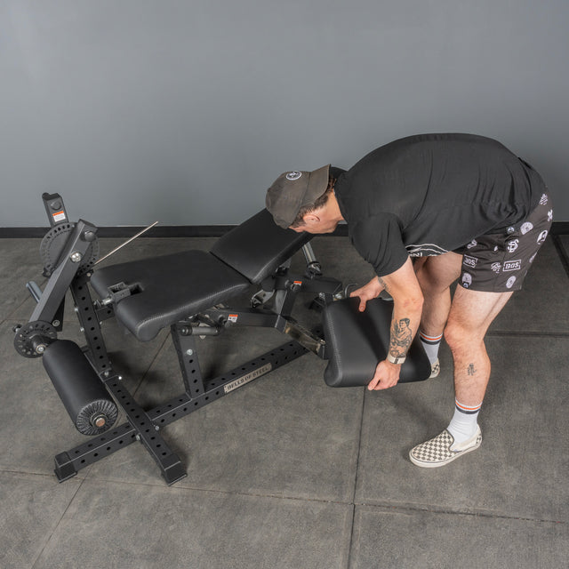 A person adjusts the leg extension on a Bells of Steel Legacy Leg Extension / Hamstring Curl Machine - Plate Loaded in a home gym with gray walls and flooring.
