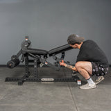 A person in casual clothes adjusts a black workout bench next to the Bells of Steel Legacy Leg Extension / Hamstring Curl Machine - Plate Loaded in a gym with gray walls and floors.