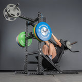 Wearing athletic gear, a person uses the Bells of Steel Pandemonium Squat – 3-in-1 Pendulum Squat / Calf Press / Viking Press Machine, loaded with blue, green, and silver plates, in a gym with gray walls and floor.