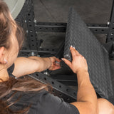 A person grips the textured, diamond-patterned metal footplate of the Bells of Steel Pandemonium Squat – a 3-in-1 Pendulum Squat / Calf Press / Viking Press machine, focusing on their hands. The scene is indoors on a gym floor.