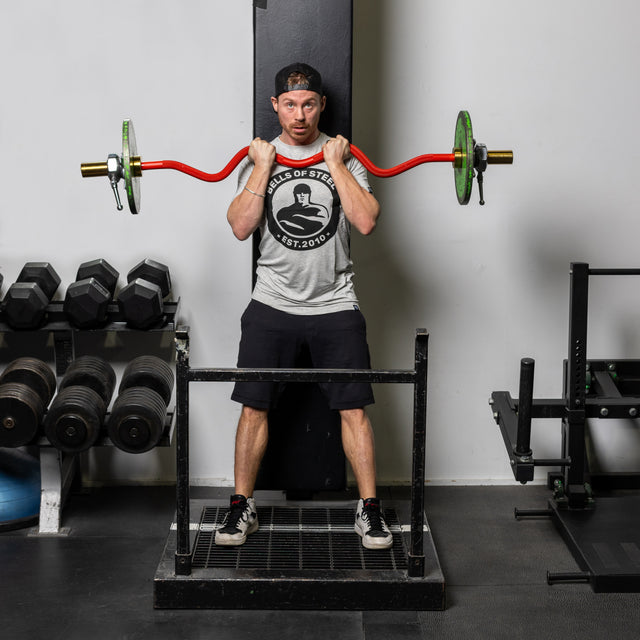 At the gym, a man stands on a platform with a Bells of Steel 100% RAW Competition Curl Bar and green weight plates on his shoulders. He sports a gray shirt, black shorts, backward cap, and sneakers. Needle bearings ensure smooth lifts amidst dumbbells and other gym equipment in the background.