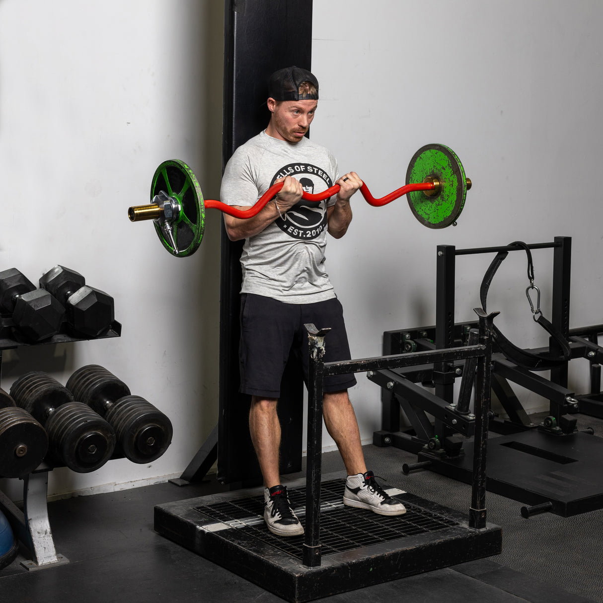 A man in a gray t-shirt and black shorts performs a barbell curl with a bright red 100% RAW Competition Curl Bar featuring green weights in the gym. Nearby, Bells of Steel dumbbells are meticulously lined up on a rack, adding to the robust atmosphere.