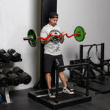 A man in a gray t-shirt and black shorts performs a barbell curl with a bright red 100% RAW Competition Curl Bar featuring green weights in the gym. Nearby, Bells of Steel dumbbells are meticulously lined up on a rack, adding to the robust atmosphere.