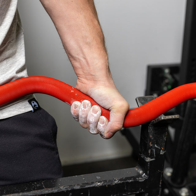 In the gym, a person grips a vibrant red, rope-like object with chalk-covered hands. They're in a light shirt and dark pants, standing near the Bells of Steel 100% RAW Competition Curl Bar, ready for their workout.