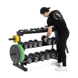 A person wearing black athletic attire organizes hex dumbbells on a Bells of Steel Residential Dumbbell Rack, part of a home gym setup with neatly arranged plate weights. Sporting a cap, the individual focuses intently on the task.