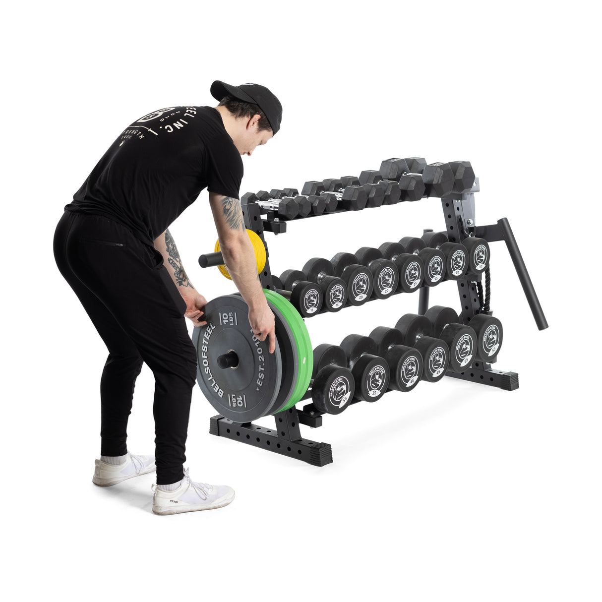 In a gym, someone wearing black and white sneakers places a weight plate on the middle shelf of the Bells of Steel Residential Dumbbell Rack, surrounded by hex dumbbells.