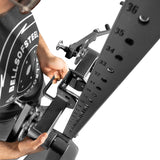 Wearing a Bells of Steel shirt, a person adjusts the Shoulder Boulder/Chest Fly Attachment, aligning its pin with the numbered holes. The overhead view highlights the mechanism designed for versatile upper body exercises.