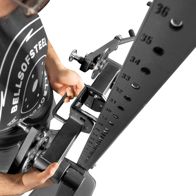 Wearing a Bells of Steel shirt, a person adjusts the Shoulder Boulder/Chest Fly Attachment, aligning its pin with the numbered holes. The overhead view highlights the mechanism designed for versatile upper body exercises.